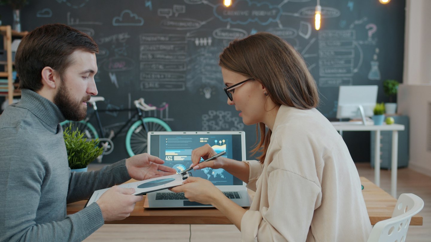 Professionals collaborate over laptop and charts at a wooden desk.